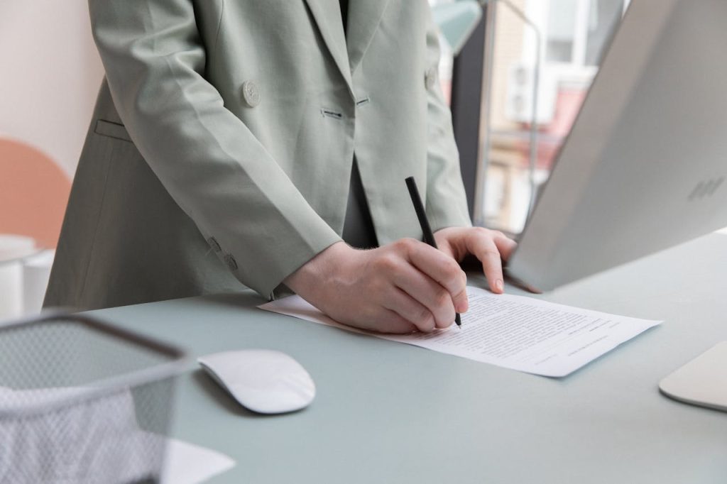 Unrecognizable worker standing at table with computer while taking notes in document while working in office